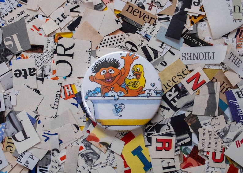May include: A round button featuring a cartoon image of a character in a bathtub with a rubber duck. The button is set against a background of cut-up newspaper clippings, with various words and letters visible.