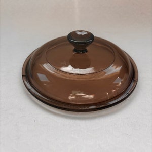 May include: A brown glass lid with a knob on top. The lid has a raised rim and the words "Anchor Hocking" are printed on the bottom.