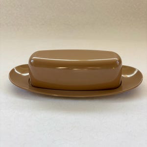May include: A brown butter dish with a lid. The dish is oval-shaped with a matching lid. The butter dish is made of ceramic or porcelain. The dish is a solid, warm brown color.