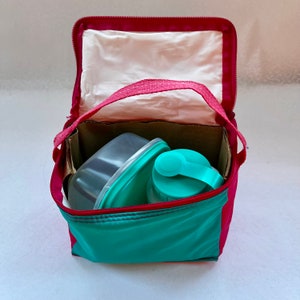 May include: A red and teal insulated lunch bag with a zipper closure. The bag is open and contains a clear plastic container and a teal water bottle with a blue lid.