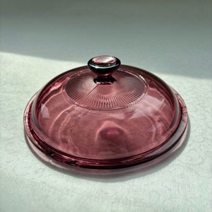 Puede incluir: Una tapa de vidrio morado con un pomo en la parte superior. La tapa tiene un diseño texturizado y acanalado.