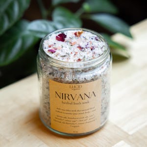 May include: A clear glass jar filled with Nirvana herbal bath soak. The jar has a tan label with the word "NIRVANA" in large letters. The bath soak is a mix of salts and dried flower petals. The jar is sitting on a light brown wooden surface.