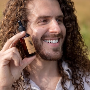May include: A small amber glass bottle with a black dropper cap, featuring a tan rectangular label with a black botanical illustration and the text SILK SERUM hair + scalp. The bottle is held upright, showcasing the dark glass and clear label.