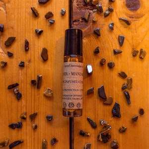 May include: A clear glass essential oil roller bottle with a black cap, labeled with text. The bottle is surrounded by small, brown and tan gemstones on a wooden surface. The text on the label includes "lucid lavender" and "DER + MAND".