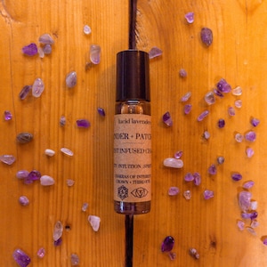 May include: A small brown glass bottle with a black rollerball top. The bottle is labeled "lucid lavender", "lavender + patchouli", "crystal infused oil", "intuition, spirit", "chakras of interest", "crown - third eye". The bottle is surrounded by small purple and clear crystals on a wooden surface.