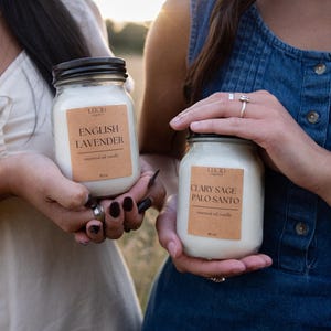 May include: Two glass jar candles with black lids and tan labels. One label reads Lucid Organics English Lavender essential oil candle 16 oz. The other label reads Lucid Organics Clary Sage Palo Santo essential oil candle 16 oz. Both jars contain white wax.