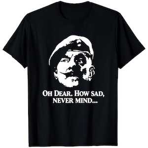 Puede incluir: Camiseta negra con un gráfico blanco de un hombre con boina y bigote. El texto "Oh Dear. How Sad, Never Mind..." está impreso debajo. Una camiseta humorística.