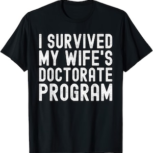 May include: Black t-shirt with white text that reads "I SURVIVED MY WIFE'S DOCTORATE PROGRAM". The text is in a bold, sans-serif font. The shirt is a crew neck style.