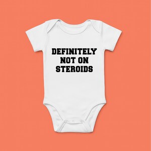 Puede incluir: Body de bebé blanco con el texto "DEFINITELY NOT ON STEROIDS" en negrita y letras mayúsculas negras. El body tiene mangas cortas y cierre de broche en la parte inferior. El fondo es de color naranja.