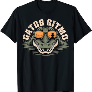 May include: Black t-shirt featuring a cartoon alligator wearing orange sunglasses. The alligator is in water and the text "GATOR GITMO" is above the alligator in an arch shape. The sunglasses reflect a structure.