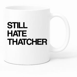 Puede incluir: Taza de cerámica blanca con el texto "STILL HATE THATCHER" en letras negras en negrita. La taza tiene un asa curva y un diseño sencillo y limpio. El texto está centrado en la superficie de la taza.