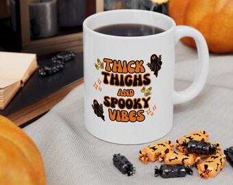 May include: White ceramic coffee mug with black text that reads "Thick Thighs and Spooky Vibes". The mug has a black ghost and orange and white candy corn graphics. The mug is sitting on a white and grey surface with orange and black candy.