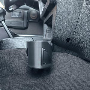 May include: Black cup holder installed in a vehicle's interior. The cup holder is cylindrical with a flat base and is mounted near the dashboard. The interior is black with a textured surface. The logo "WILLOW DESIGN HOUSE" is visible.