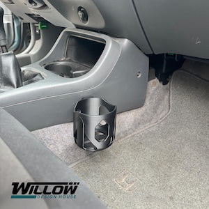 May include: Black car cup holder with a geometric design, placed near the center console of a vehicle. The cup holder is made of a dark material and is designed to fit in the car's interior. The Willow Design House logo is visible at the bottom.