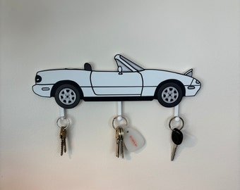 Puede incluir: Porta llaves blanco con forma de coche descapotable con tres ganchos. El coche tiene contornos negros y ruedas negras. Hay tres llaves colgando de los ganchos.