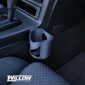 May include: A gray car cup holder, mounted near the center console. The cup holder is made of plastic and has a curved design with a cutout for a handle. The logo "Willow Design House" is visible at the bottom.