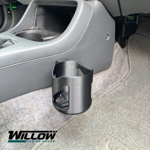 Puede incluir: Portavasos negro para coche con un diseño curvo, montado en un salpicadero gris. El portavasos tiene un recorte en el lateral y está situado cerca de la consola central. El texto "WILLOW DESIGN HOUSE" está en la parte inferior.