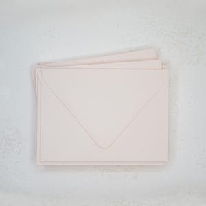 Puede incluir: Una pila de sobres de color rosa pálido dispuestos sobre una superficie gris claro. Los sobres son rectangulares con un diseño de solapa puntiaguda clásico. El papel tiene una textura suave y un color suave y apagado.
