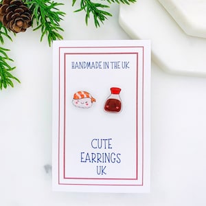 May include: A white card with the text "HANDMADE IN THE UK" and "CUTE EARRINGS UK". The card shows a pair of earrings, one is a cartoon sushi with a smiling face and the other is a cartoon bottle of soy sauce with a smiling face.