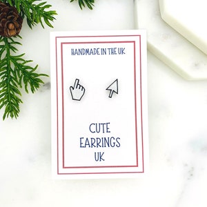May include: A white card with black text that reads "Handmade in the UK" and "Cute Earrings UK". The card features two white computer mouse cursor icons with black outlines. The icons are a pointing hand and a standard arrow.