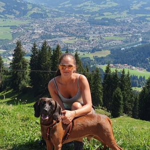 May include: A woman wearing a vest top and sunglasses is sitting on a path with a brown and black dog. The dog is wearing a red and black harness. The background is a mountain range with a town in the valley. The text "Hundalltag" is written in white on a green background with a red heart.