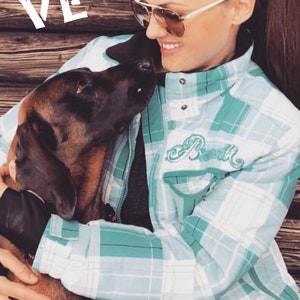 May include: A woman wearing a light blue and white checked shirt with the word "Rustik" embroidered on the left side of the chest. She is holding a brown and black dog in her arms. The dog is looking up at the woman. The woman is wearing sunglasses and has her hair tied back in a ponytail.