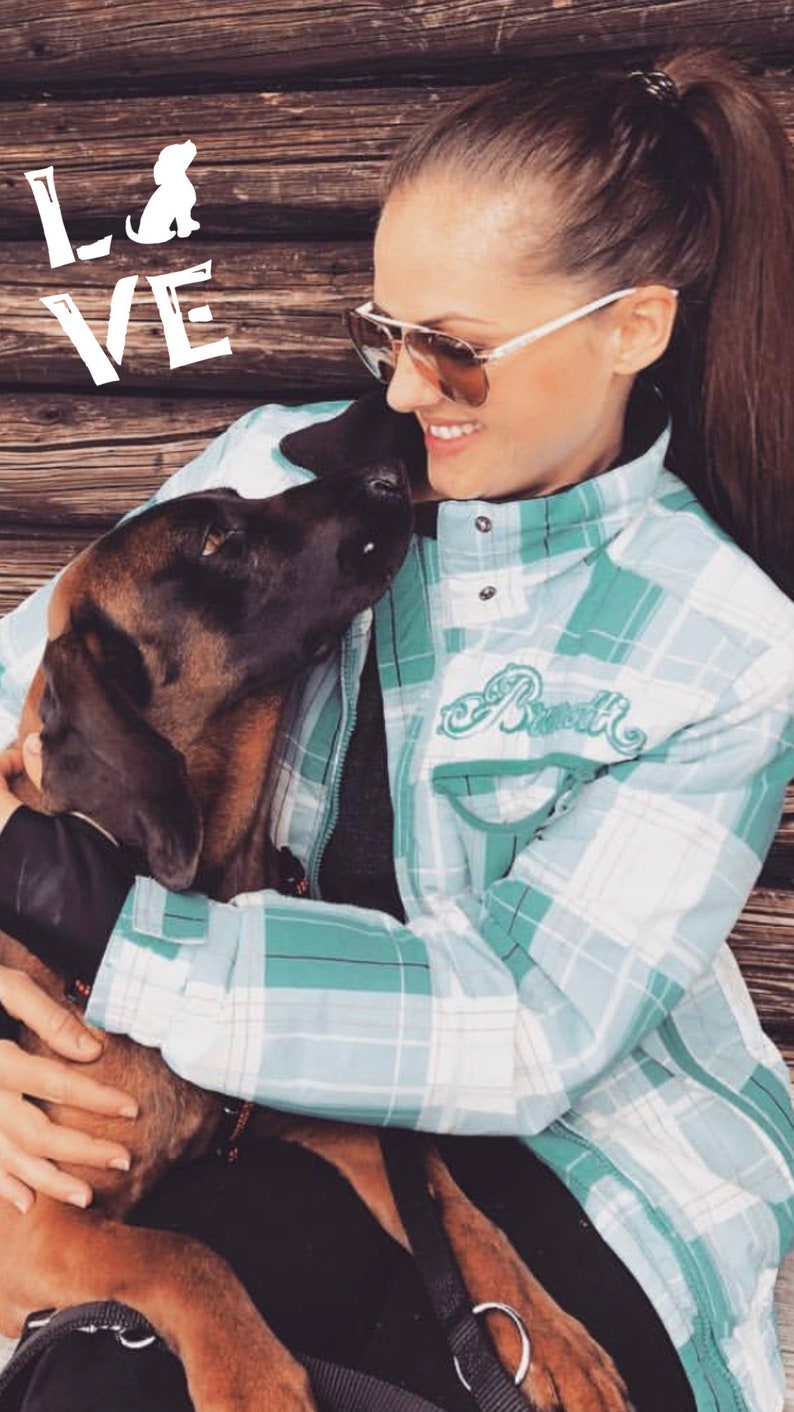 May include: A woman wearing a light blue and white checked shirt with the word "Rustik" embroidered on the left side of the chest. She is holding a brown and black dog in her arms. The dog is looking up at the woman. The woman is wearing sunglasses and has her hair tied back in a ponytail.