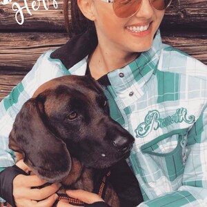 May include: A woman wearing sunglasses and a green and white checked shirt is holding a brown and black dog. The shirt has the word "Brammott" embroidered on the front. The woman is smiling and the dog is looking at the camera.