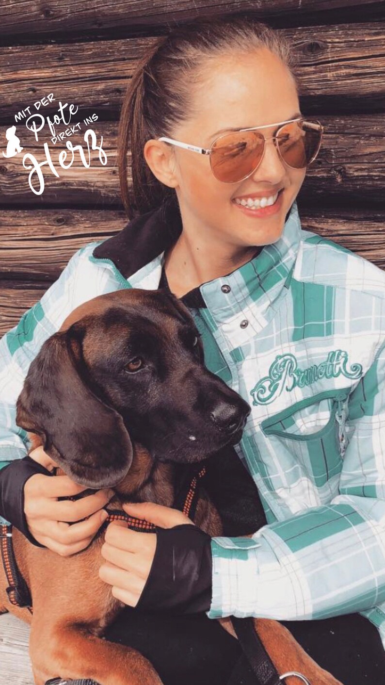 May include: A woman wearing sunglasses and a green and white checked shirt is holding a brown and black dog. The shirt has the word "Brammott" embroidered on the front. The woman is smiling and the dog is looking at the camera.