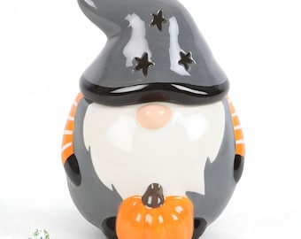 May include: A ceramic gnome figurine with a grey hat featuring star cutouts and a black brim. The gnome has a white beard, a pink nose, and is holding an orange pumpkin. Orange and white stripes accent the sides. The figurine is a Halloween decoration.