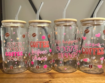 May include: Four clear glass coffee tumblers with bamboo lids and glass straws. Each tumbler has a different coffee-themed phrase in pink and orange, along with coffee bean and star decorations. The phrases include "Anti Social Coffee Club".