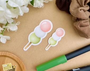May include: Two dango stickers, a Japanese sweet treat, on a brown background. The dango are white, green, and pink. The stickers are perfect for adding a touch of Japanese culture to your planner, laptop, or water bottle.