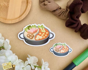 May include: Two illustrated stickers of a Yin Yang hot pot with various ingredients, including tofu, mushrooms, and vegetables. The stickers are on a brown surface with white flowers and a green pen.
