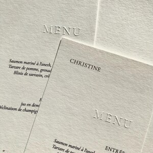 Może przedstawiać: Zbliżenie kilku białych papierowych menu z wytłoczonym tekstem. Słowo "MENU" jest wyraźnie wyświetlane na każdym z nich. Jedno menu jest oznaczone "ANNE", drugie "CHRISTINE", a trzecie zawiera francuskie menu z nagłówkiem "ENTRÉE".
