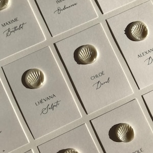 Peut inclure: Gros plan sur d'élégants cartons de table avec des noms en écriture cursive. Chaque carte est ornée d'une décoration en forme de coquillage. Les cartes sont disposées avec soin, suggérant un mariage ou un événement spécial.