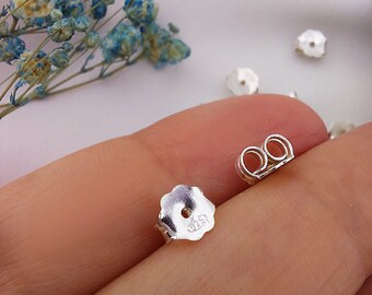 May include: Silver earring backs with a butterfly design and a 925 stamp. The backs are shown on a finger.