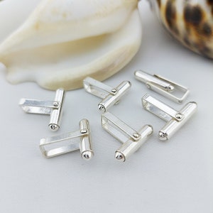 May include: Six shiny silver cufflinks with a rectangular and cylindrical design. The cufflinks are arranged on a white surface, with a seashell in the background. The cufflinks have a small, round end and a rectangular clasp.