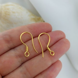 May include: Two gold-colored earring hooks are displayed. Each hook has a curved design with a looped end and a coiled wire detail. The hooks are held in a hand, suggesting their small size and use in jewelry making.
