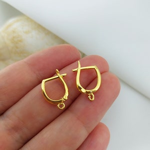 May include: Two gold-coloured earring findings. Each finding has a curved, D-shaped design with a hinged top and a small loop at the bottom. The findings are made of a shiny, reflective metal.