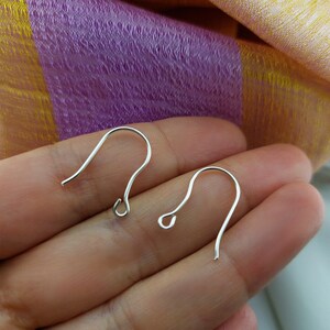 May include: Two silver-colored earring hooks are displayed against a background of purple and yellow fabric. The hooks are curved with a small loop at the bottom, designed for attaching earrings. The hooks are made of a shiny metal.