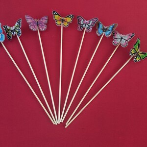 May include: A set of eight wooden sticks with colorful butterfly decorations. The butterflies are made of iridescent material and have different patterns and colors.