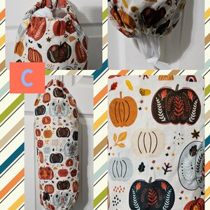 May include: A white fabric bag with a pumpkin pattern in orange, brown, and blue. The bag is designed to hold plastic grocery bags and has a drawstring closure. The bag is hanging on a hook.