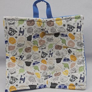 May include: A square, quilted pot holder with a blue handle and trim. The fabric features a repeating pattern of Star Wars characters and symbols in various colors on a white background. The pot holder is designed to protect hands from hot cookware.