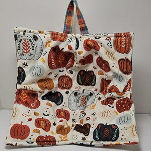 May include: A white quilted hot pad with a pumpkin pattern in shades of orange, brown, and blue. The hot pad has a plaid handle.