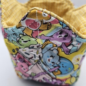 May include: A colorful, quilted bowl cozy featuring a pattern of cartoon bears in various colors. The interior is a solid yellow, and the exterior has a vibrant, multi-colored design. The cozy is designed to hold a bowl of food and protect hands from heat.