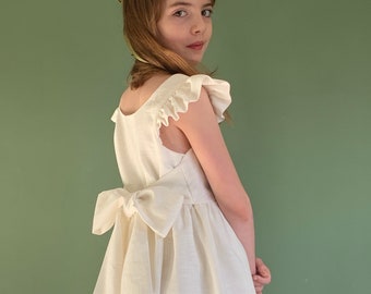 Op de afbeelding: Een jong meisje draagt een witte linnen jurk met een grote strik op de rug. De jurk heeft geruite mouwen en een eenvoudig, elegant ontwerp. Ze draagt een groene bloemenkrans in haar haar.