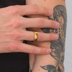 Könnte beinhalten: Ein goldener Bandring mit quadratischer Form. Der Ring wird am Ringfinger einer Hand getragen.