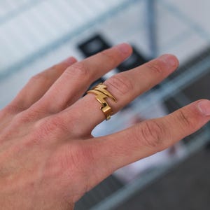 Op de afbeelding: Een gouden ring met een geometrisch ontwerp. De ring wordt gedragen aan de middelvinger van een hand.