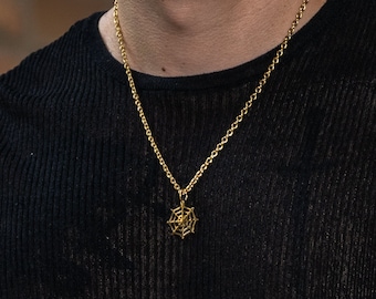 Pode incluir: Colar dourado com um pendente de teia de aranha. O pendente apresenta um design detalhado de teia. O colar é usado com uma camisa preta de manga comprida. Uma pulseira dourada também é visível no pulso.