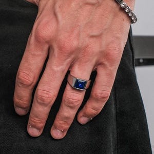 Könnte beinhalten: Ein silbernes Armband mit klaren, quadratisch geschliffenen Steinen und ein silberner Ring mit einem quadratischen, dunkelblauen Stein. Der Ring befindet sich am Ringfinger. Die Person trägt eine schwarze Jeans und ein schwarzes Hemd.